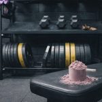 En skopa proteinpulver på en träningsbänk i ett gym med vikter i bakgrunden.