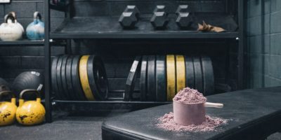 En skopa proteinpulver på en träningsbänk i ett gym med vikter i bakgrunden.